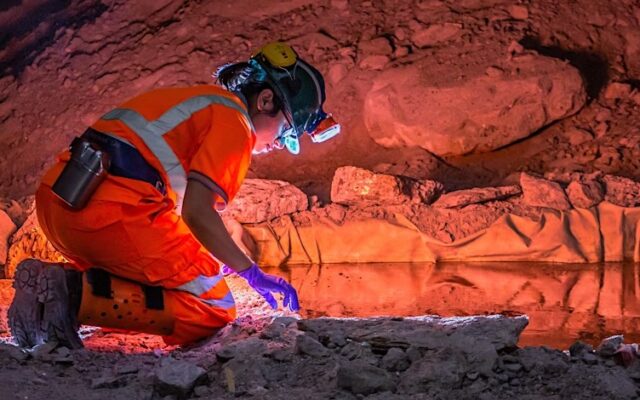 Scientist in safety gear kneeling in an underground cave at Boulby Underground Laboratory, examining rocks near a reflective pool under orange lighting.