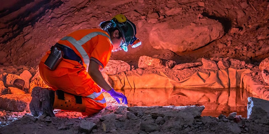 Scientist in safety gear kneeling in an underground cave at Boulby Underground Laboratory, examining rocks near a reflective pool under orange lighting.