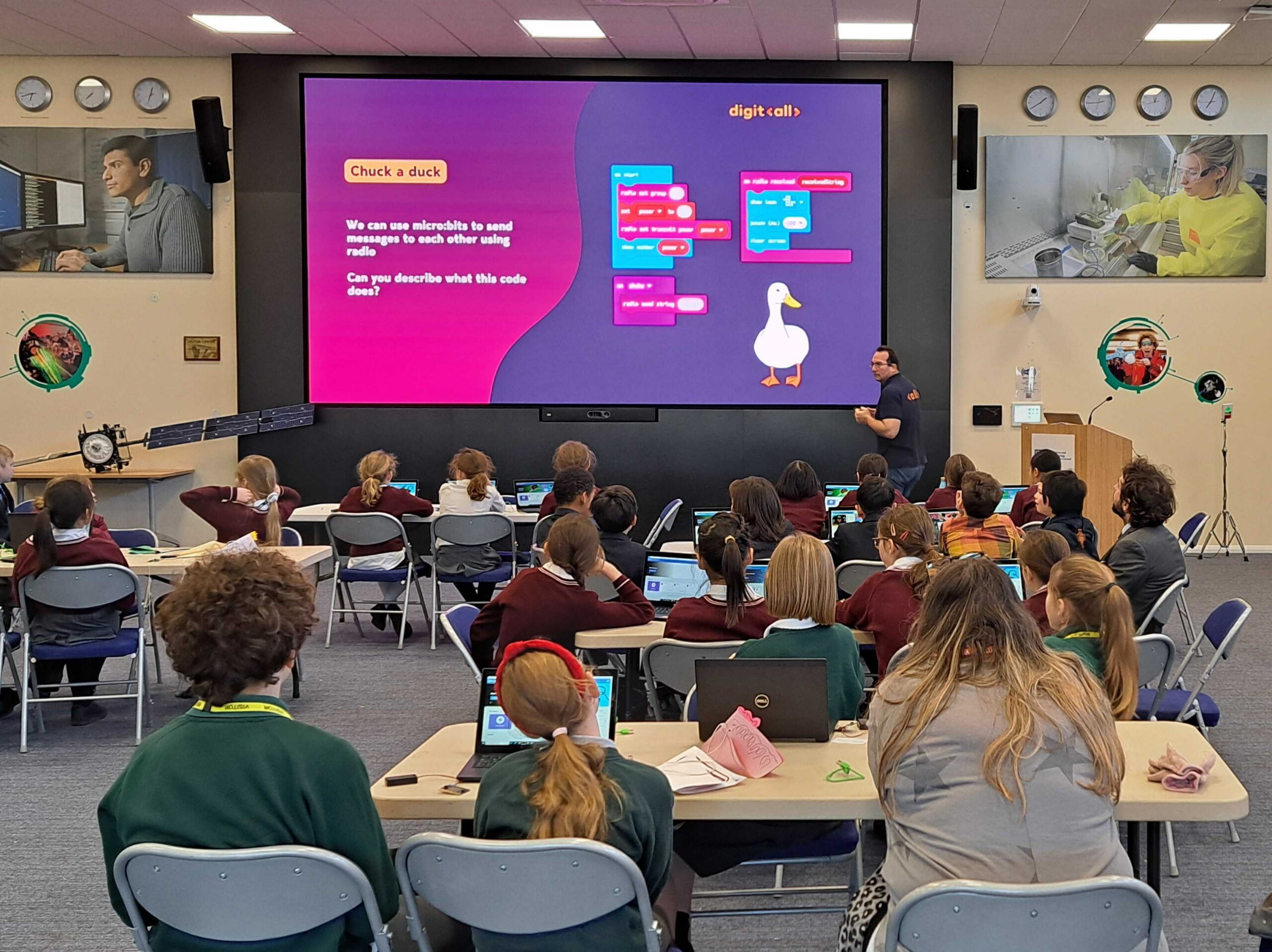 a pink and purple presentation slide can be seen on a large screen, students sit in groups around tables watching the presentation.