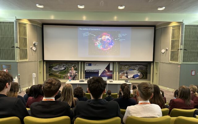 View from the back of a lecture theatre, behind a row of students looking to a screen with a slide about Cassiopeia