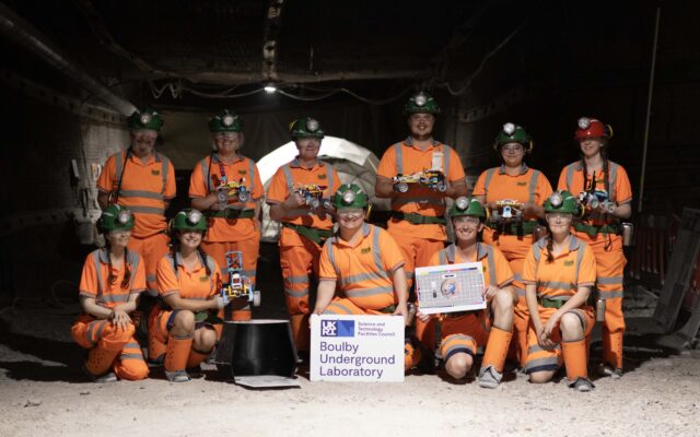 The Boulby Underground Laboratory team in a mine tunnel. Dressed in bright orange safety gear holding lego robots.