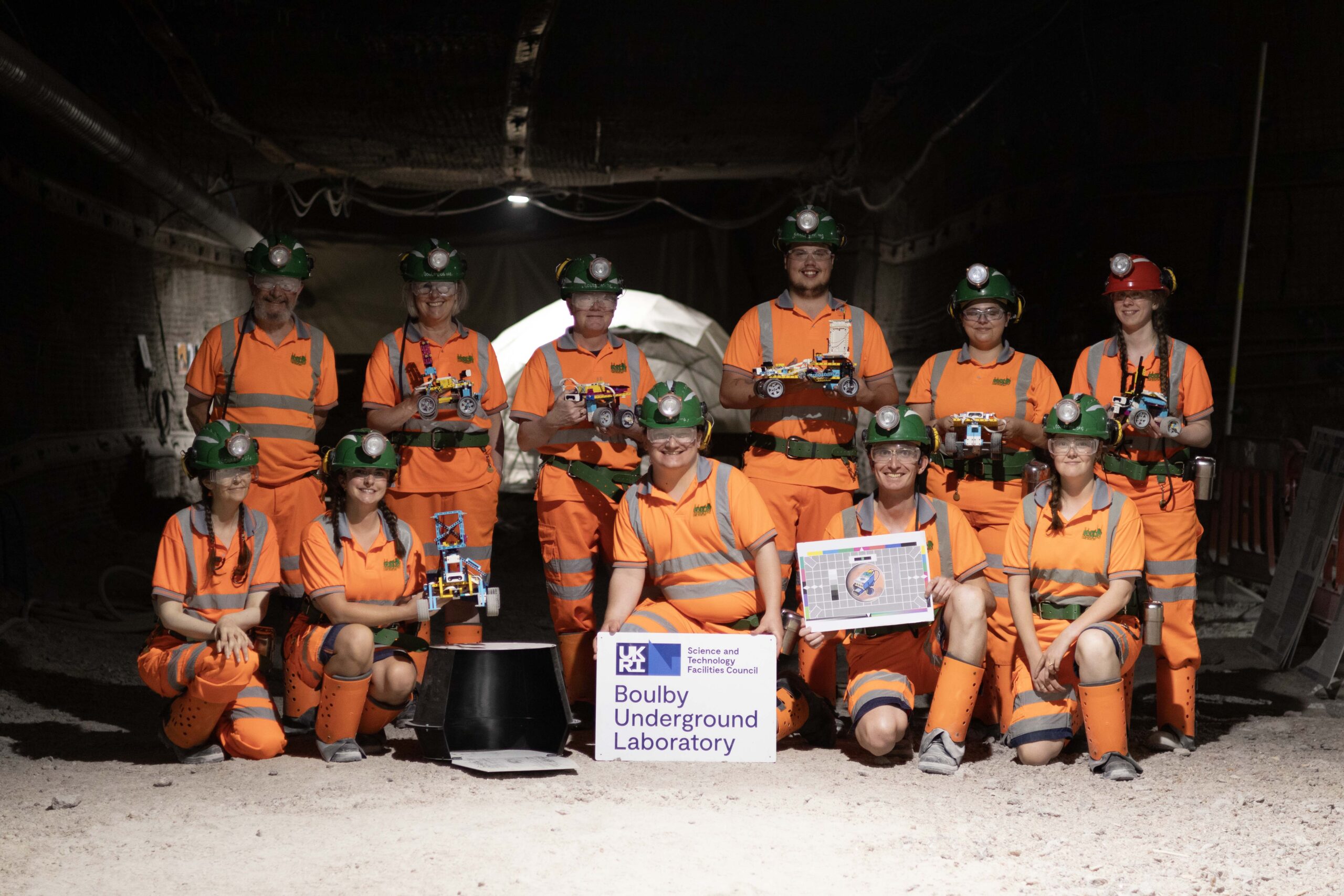 The Boulby Underground Laboratory team in a mine tunnel. Dressed in bright orange safety gear holding lego robots.