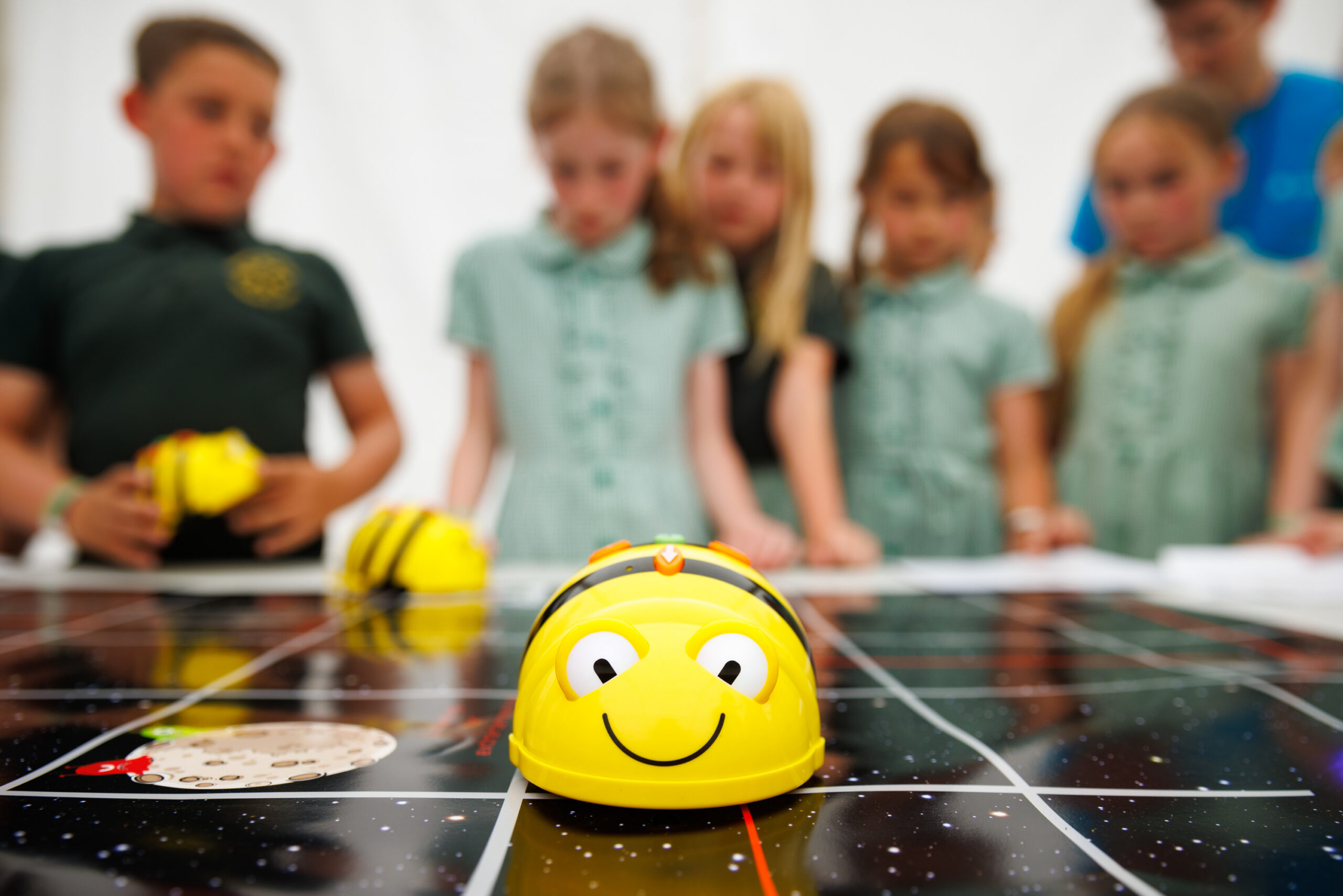 a yellow bee robot is on a black map following a red line. In the blurred background, children can be seen watching the beebot.
