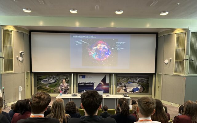 Students in a lecture theatre look at a presentation on a large screen at the front of the hall.