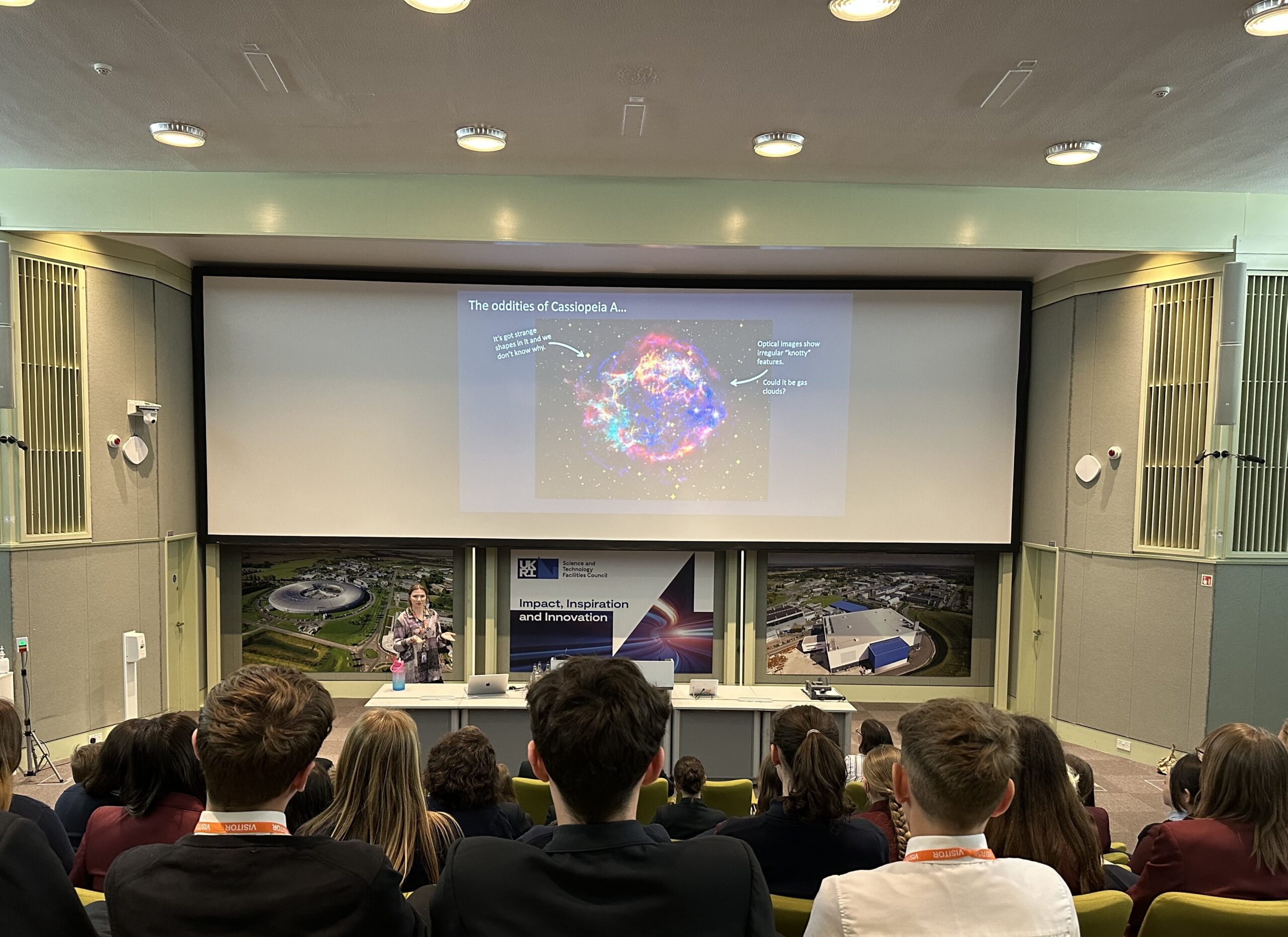 Students in a lecture theatre look at a presentation on a large screen at the front of the hall.