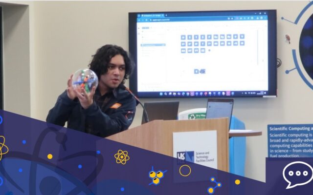 A person stands at a wooden lectern and holds a clear sphere with blue and orange sections inside. Behind them a monitor screen on the wall shows a table of blue squares