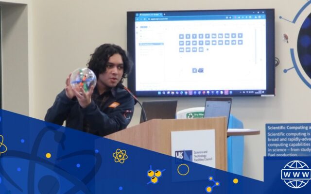 A person stands at a wooden lectern and holds a clear sphere with blue and orange sections inside. Behind them a monitor screen on the wall shows a table of blue squares