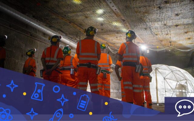 A group of people in orange hi-visibility clothing with green helmets and yellow ear defenders stand in one of the Boulby underground laboratory tunnels, a plastic habitat dome can be seen in the background