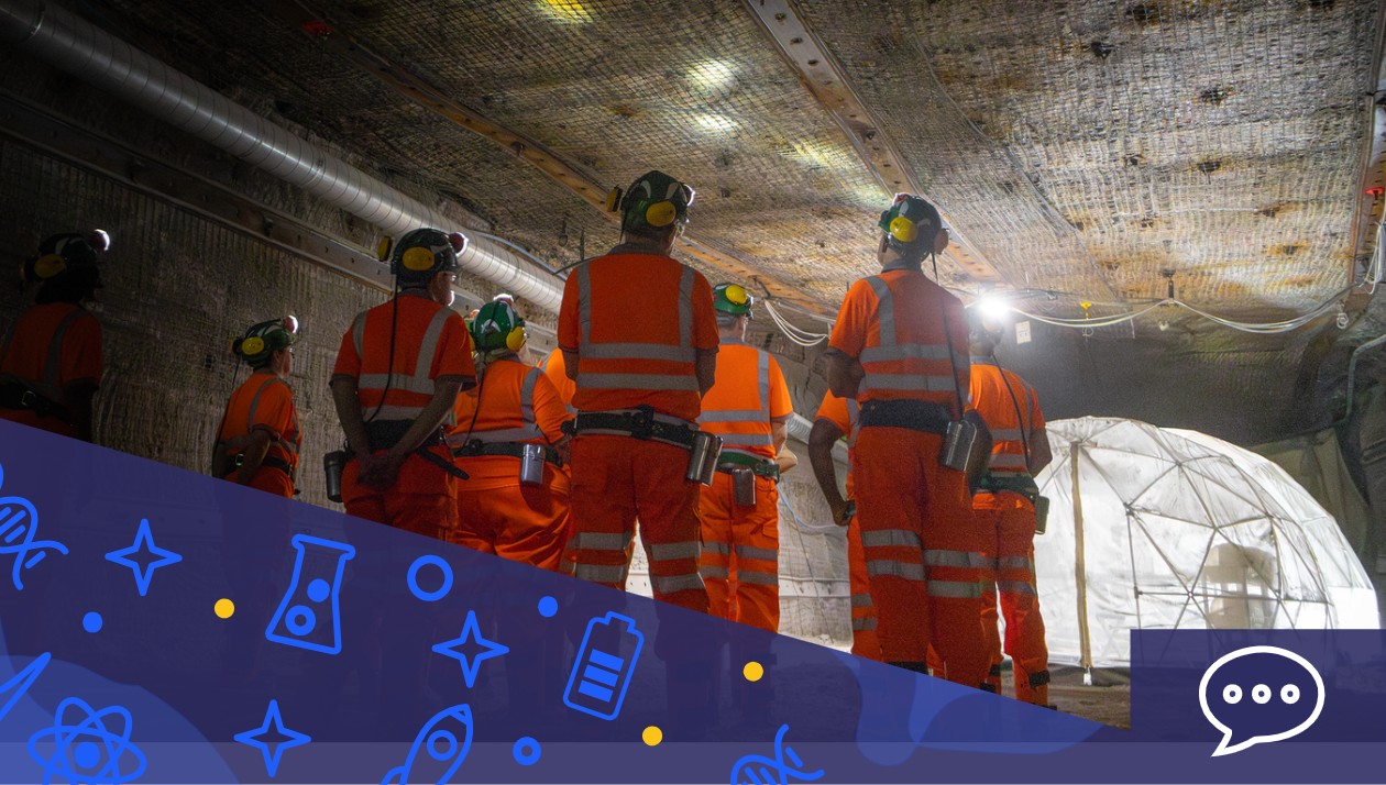 A group of people in orange hi-visibility clothing with green helmets and yellow ear defenders stand in one of the Boulby underground laboratory tunnels, a plastic habitat dome can be seen in the background